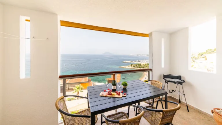 Balcón con mesa, sillas de mimbre, botella de vino, vista al mar y playa. Parrilla en una esquina. Ambiente luminoso.