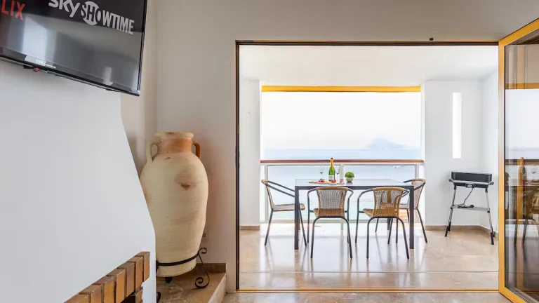 Televisor en pared blanca, jarrón grande, terraza con mesa y sillas; vistas al mar. Parrilla a la derecha, botella y copas en la mesa.