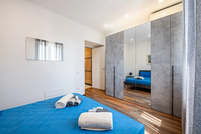 Habitación moderna con cama cubierta por manta azul, toallas dobladas, armario de espejos y suelo de madera. Luz natural.