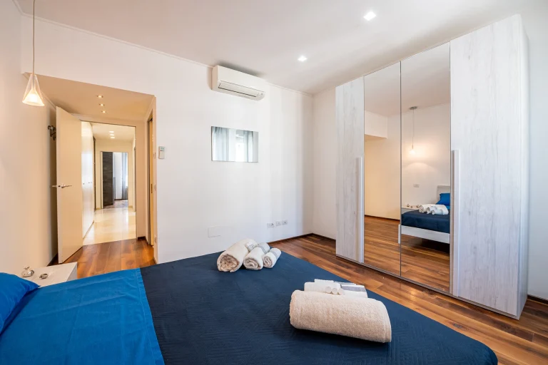 Habitación moderna con cama de sábanas azules, armario con espejo, suelo de madera y luces empotradas en el techo.