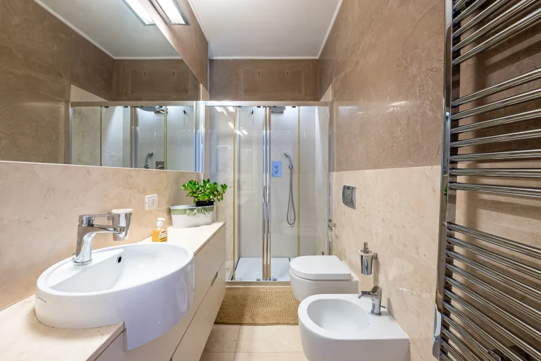 Baño moderno con lavabo blanco ovalado, ducha con puerta de vidrio, inodoro, bidé y toallero de acero inoxidable. Decoración minimalista.