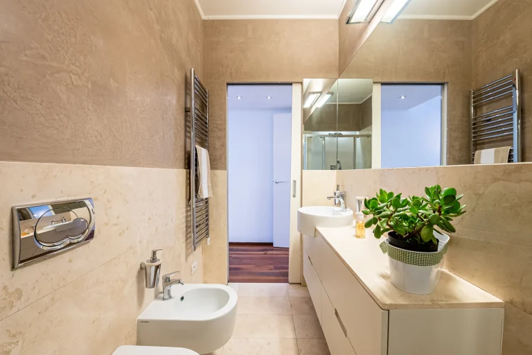 Baño moderno con paredes beige, lavabo blanco, espejo grande, planta en encimera y suelo de baldosas claras.