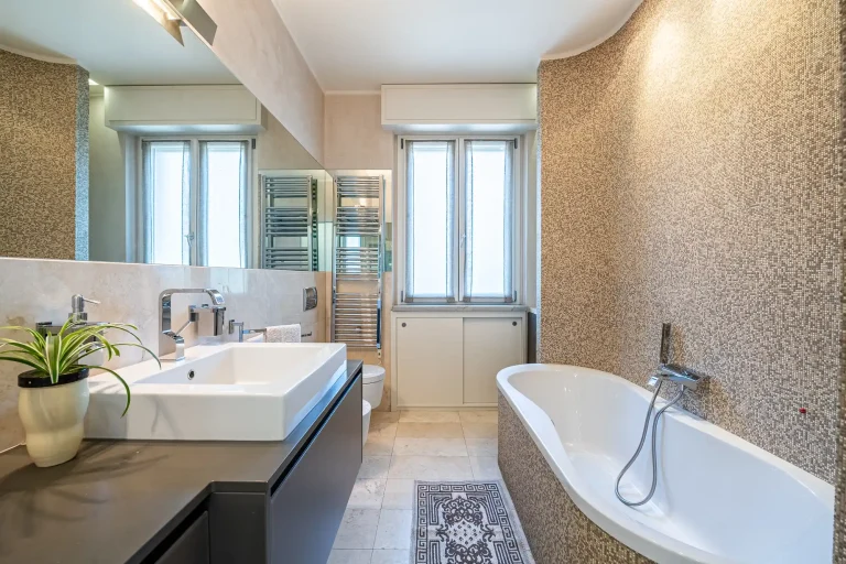 Baño moderno con lavabo blanco, planta decorativa, bañera ovalada, ventanas grandes y toallas blancas. Suelo claro y pared con mosaico.