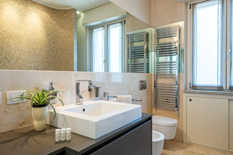 Baño moderno con paredes claras, lavabo blanco, grifería cromada, planta decorativa, ventana y toalla cerca del inodoro.