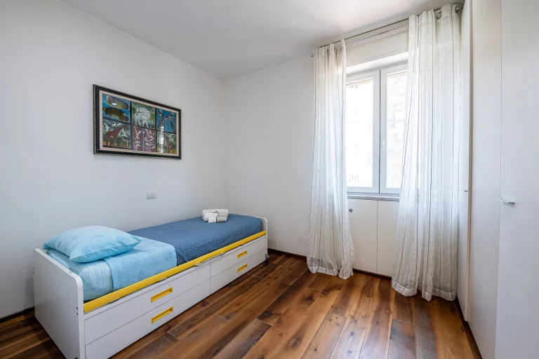 Habitación minimalista con cama individual, cubrecama azul, almohada celeste, piso de madera, cortinas blancas y cuadro colorido en la pared.