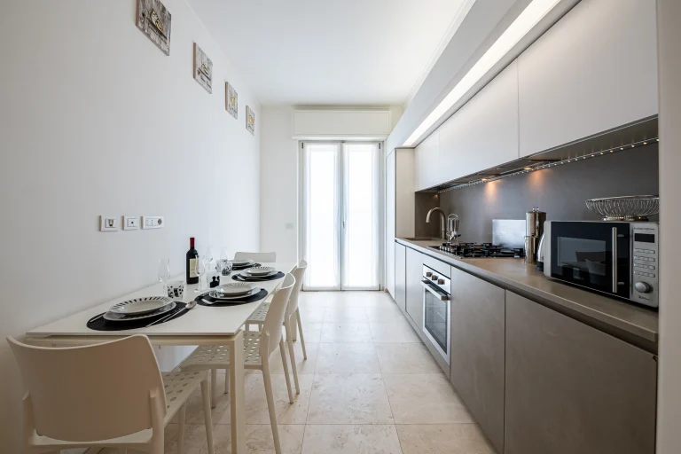 Cocina moderna con paredes blancas, encimera gris, microondas, mesa con cuatro sillas y vajilla, luz natural entrando por ventanales.