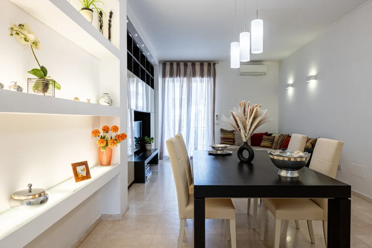 Comedor moderno con mesa oscura, sillas beige y lámparas colgantes. Estantería decorada con plantas y flores naranjas. Cortinas blancas.