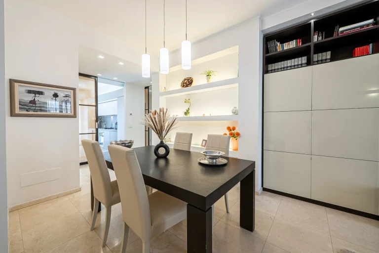 Comedor moderno con mesa negra, sillas beige, estanterías blancas con plantas y adornos. Lámparas colgantes iluminan el espacio.