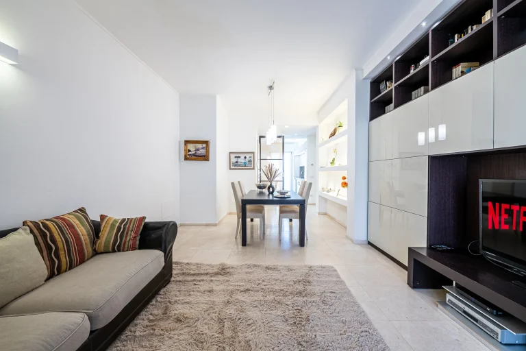 Sala moderna con sofá y alfombra beige, comedor al fondo. TV con logo de Netflix a la derecha. Decoración minimalista.