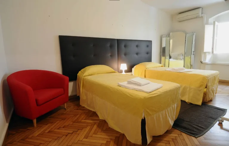 Habitación con dos camas individuales amarillas, cabecera negra, silla roja y ventana abierta. Piso de parquet y lámpara de mesita.