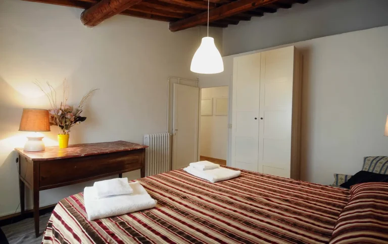 Habitación con cama de rayas marrones y rojas, escritorio con lámpara y jarrón, armario blanco, techo con vigas de madera.