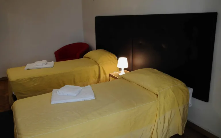 Dos camas individuales cubiertas con sábanas amarillas, lámpara encendida en mesa de noche, silla roja en el rincón, pared clara de fondo.