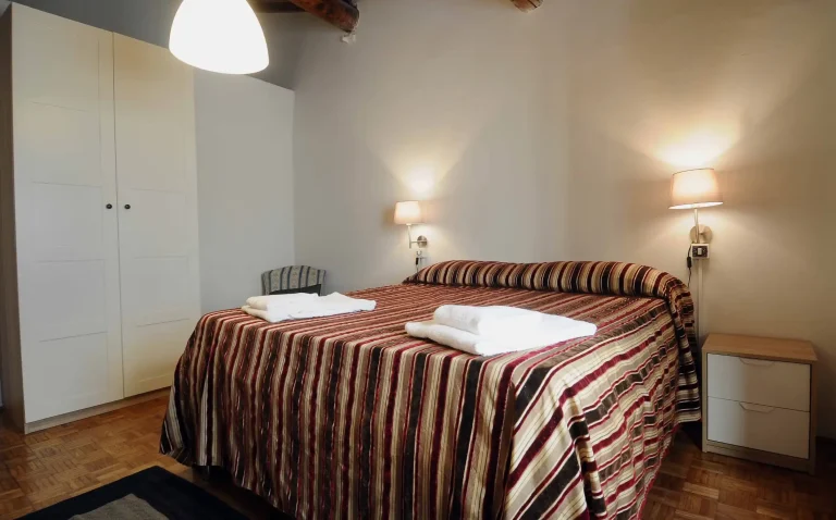 Habitación con cama grande, colcha de rayas marrones, luces de pared, armario blanco y suelo de madera.