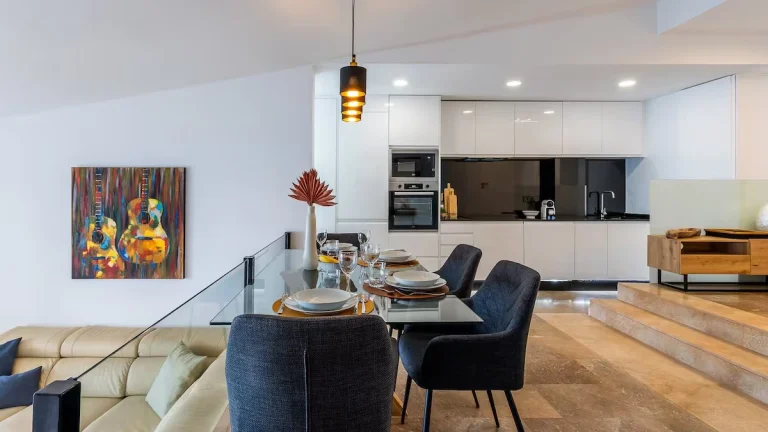 Cocina moderna con gabinetes blancos, mesa de vidrio con sillas negras, y cuadro colorido de guitarras en la pared.