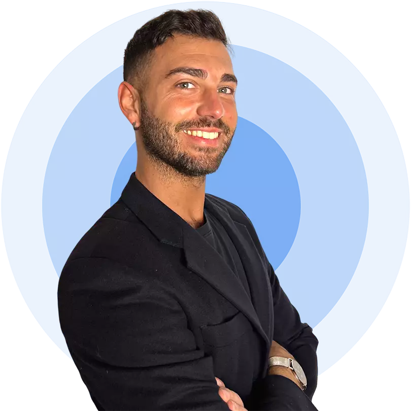 Hombre de barba y sonrisa, vestido con blazer negro y camiseta oscura. Fondo circular azul con líneas concéntricas. Pose con brazos cruzados.