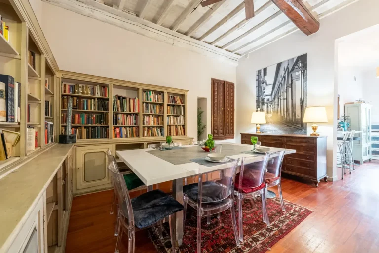 Comedor con mesa blanca, sillas transparentes, estanterías con libros, lámparas y alfombra roja. Pared con arte en blanco y negro.