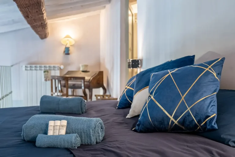 Cama con edredón azul oscuro, cojines decorativos azules con líneas doradas, toallas azules enrolladas y escritorio de madera al fondo.