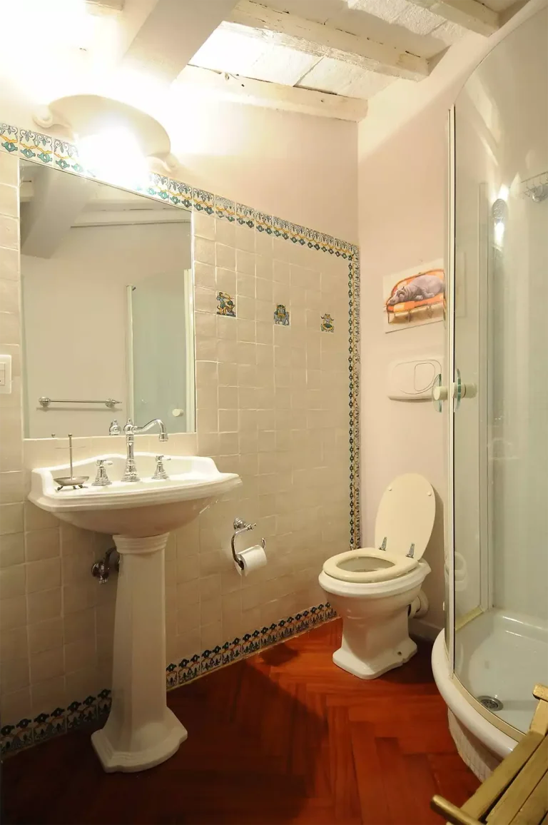 Baño con azulejos decorados, lavabo de pedestal, ducha esquinera de vidrio, inodoro y piso de madera. Iluminación en techo.