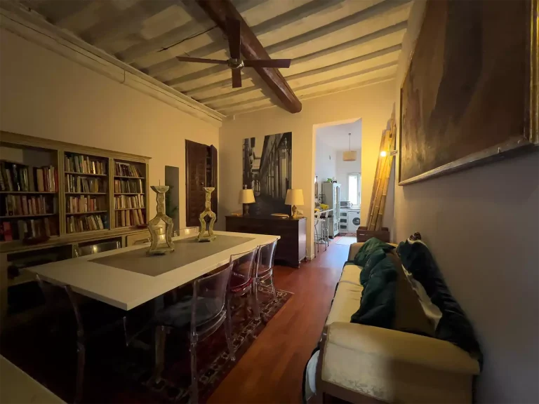 Sala de estilo clásico con mesa blanca, sillas transparentes, librero lleno, sofá con cojines y vigas de madera en el techo.