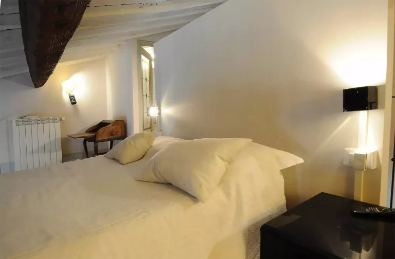 Habitación con cama doble cubierta de edredón blanco, lámparas de pared y vigas de madera en el techo inclinado.