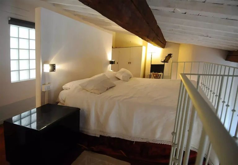 Dormitorio con cama grande cubierta por edredón blanco, ventanas de vidrio bloqueado y techo de vigas de madera expuestas.