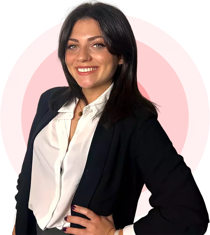 Mujer de cabello oscuro y sonrisa, vestida con blazer negro y camisa blanca. Fondo circular rosa con líneas concéntricas. Pose con una mano en la cadera.