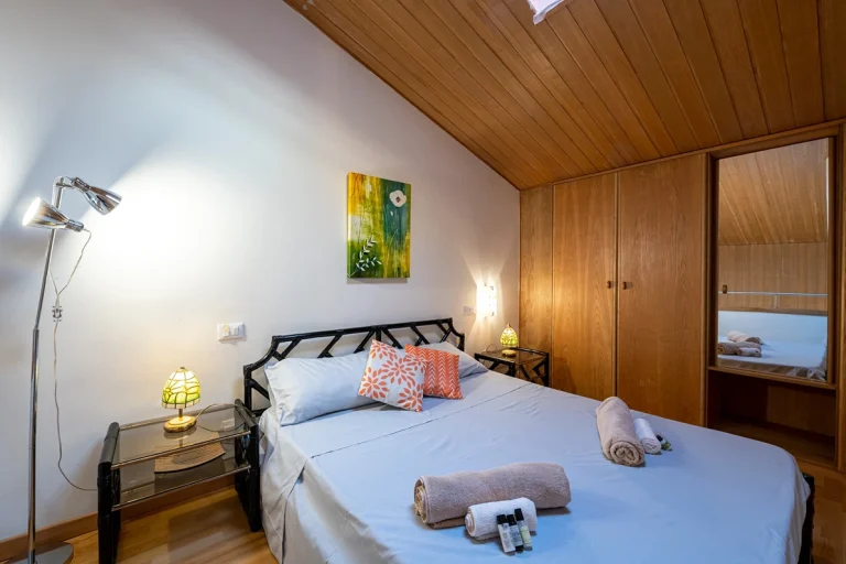 Habitación con techo de madera, cama doble con sábanas claras y cojines naranjas. Mesitas de noche con lámparas, armario con espejo y decoración moderna.