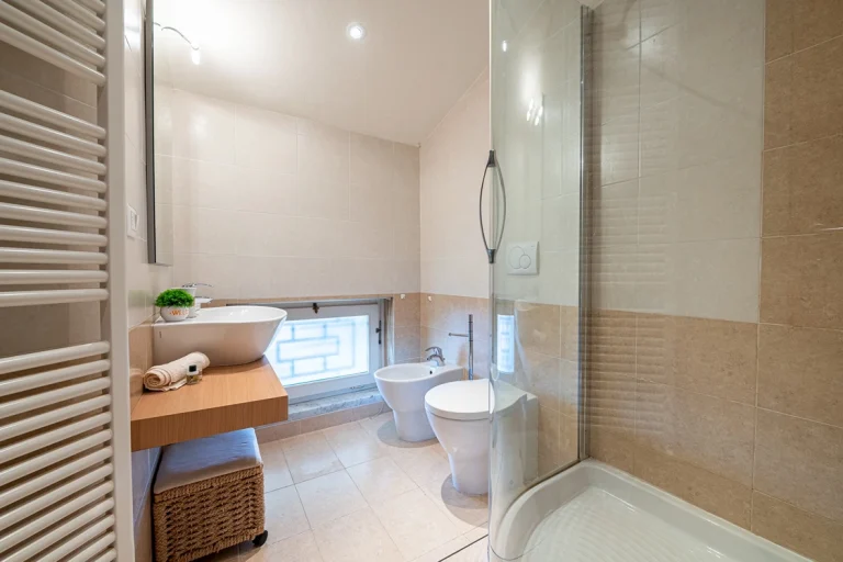 Baño moderno con ducha de cristal, lavabo sobre encimera de madera, bidé y sanitario. Paredes de azulejos beige y ventana baja con vidrio esmerilado.