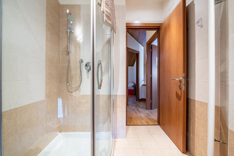 Baño con ducha de cristal y azulejos en tonos beige. Toalla colgada sobre la mampara. Puerta de madera abierta, mostrando un pasillo y una habitación.