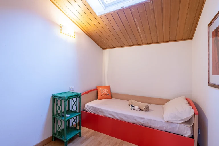 Habitación con techo de madera y ventana superior. Cama individual con cabecera roja, cojín decorativo y toallas. Estante verde y cuadro en la pared.