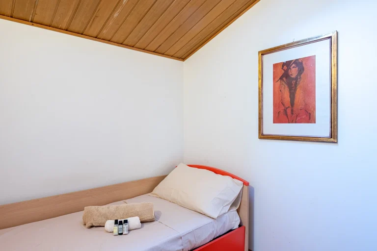 Habitación con cama individual, sábanas beige y cabecera roja. Toallas y productos de aseo sobre la cama. Cuadro con retrato en la pared.