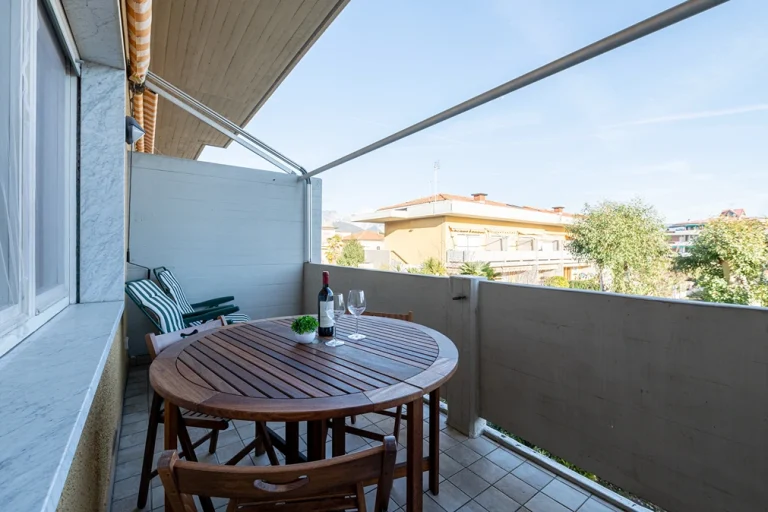 Balcón con mesa redonda de madera, sillas y una botella de vino con copas. Dos tumbonas con cojines rayados y vista a edificios y árboles bajo un cielo despejado.