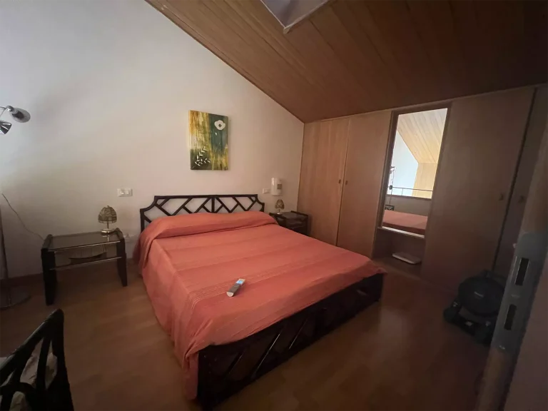 Habitación con techo de madera, cama doble con colcha naranja y cabecera negra. Mesitas de noche con lámparas, armario con espejo y luz tenue.