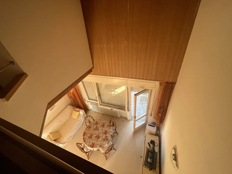 Vista superior de una sala de estar con techo de madera. Sofá blanco, mesa de comedor con mantel floral y sillas de mimbre. Ventanales amplios con cortinas naranjas.