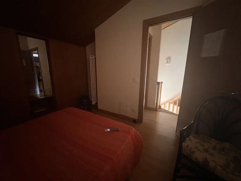 Habitación con luz tenue, cama con colcha naranja, armario con espejo y silla de mimbre. Puerta abierta con vista a una escalera de madera iluminada.