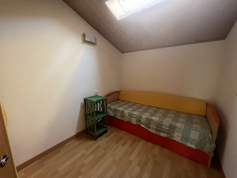 Habitación pequeña con techo inclinado, cama individual con base roja y respaldo amarillo. Estante verde de mimbre y tragaluz en el techo para iluminación natural.