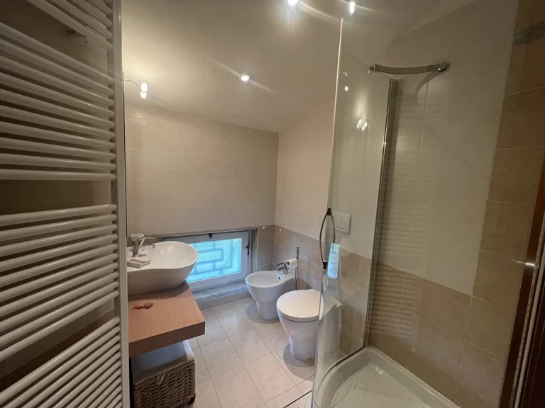 Baño moderno con ducha de cristal, inodoro y bidé. Lavabo sobre encimera y ventana baja con vidrio esmerilado. Iluminación empotrada en el techo.