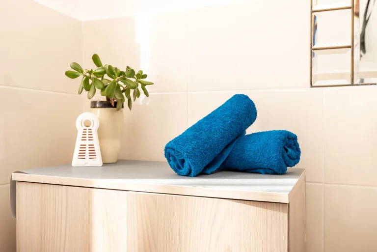 Dos toallas azules enrolladas sobre un mueble de baño de madera clara. Maceta con planta verde y ambientador blanco junto a pared de azulejos beige.