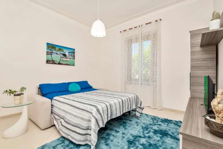 Dormitorio moderno con cama de sábanas azules y colcha a rayas. Cojín turquesa, televisor con streaming, lámpara colgante y decoración minimalista.