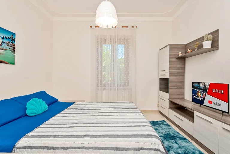 Dormitorio moderno con cama de sábanas azules y colcha a rayas. Cojín turquesa, televisor con streaming, lámpara colgante y decoración minimalista.