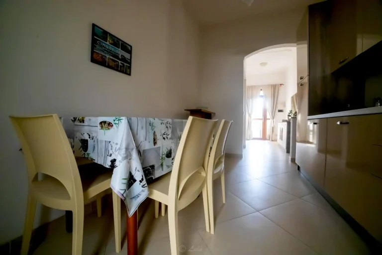 Comedor con mesa cubierta con mantel de dibujos, sillas beige, paredes claras y entrada arqueada hacia una sala iluminada con cortinas.