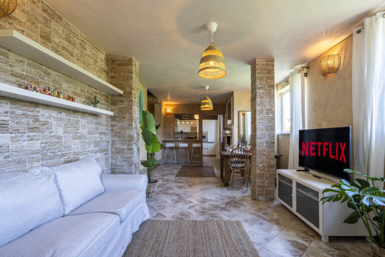 Sala con piso de mármol, sofá blanco, estanterías blancas, televisor con Netflix, lámparas de mimbre, planta y comedor al fondo.