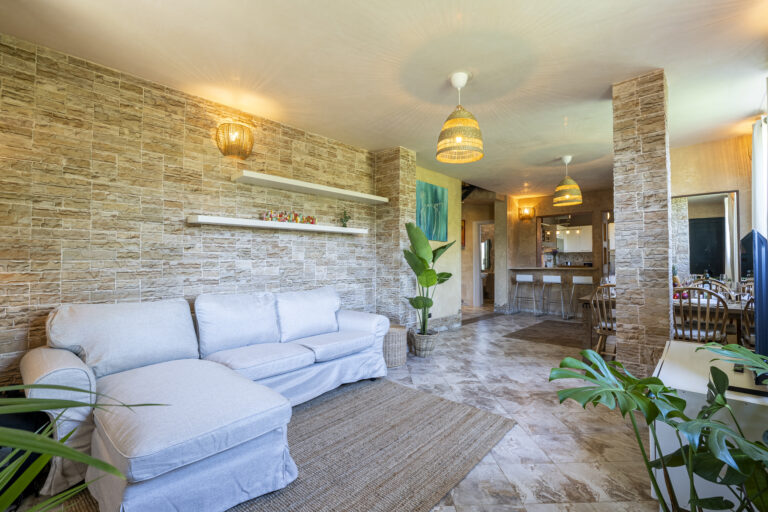 Sala de estar con sofá gris, pared de piedra, plantas en macetas, lámparas colgantes, barra con taburetes y mesa comedor al fondo.