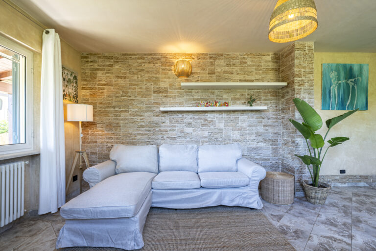 Sofá blanco frente a pared de ladrillo, lámpara de pie, planta en maceta, estante flotante y cuadro azul.
