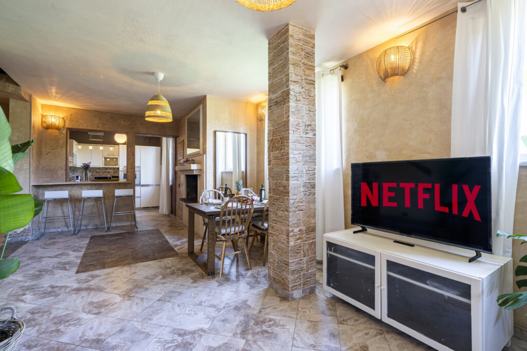 Sala de estar con TV que muestra Netflix, comedor con mesa de madera, sillas y cocina al fondo. Lámparas colgantes y paredes beige.
