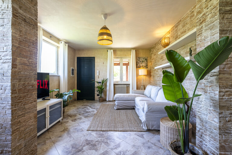 Sala moderna con paredes de piedra, sofá blanco, plantas y televisor. Lámpara colgante y alfombra beige sobre suelo de baldosas.