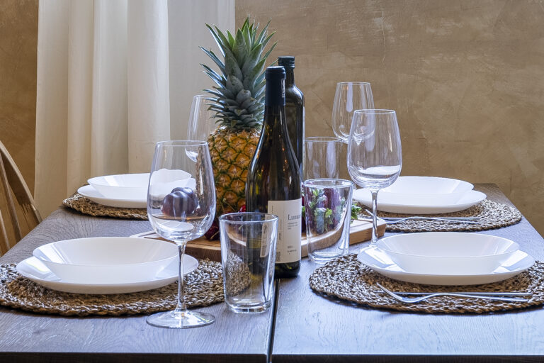 Mesa con manteles individuales tejidos, vajilla blanca, copas de vino, botellas, piña y frutas frescas como decoración central.