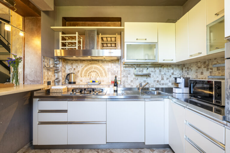 Cocina moderna con muebles blancos, encimera de acero, mosaico en la pared, horno, microondas y flores en un jarrón.