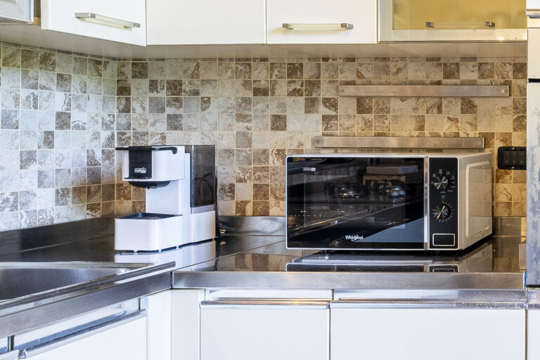 Cocina moderna con encimera de acero. Cafetera blanca a la izquierda y horno microondas plateado a la derecha; azulejos de fondo.