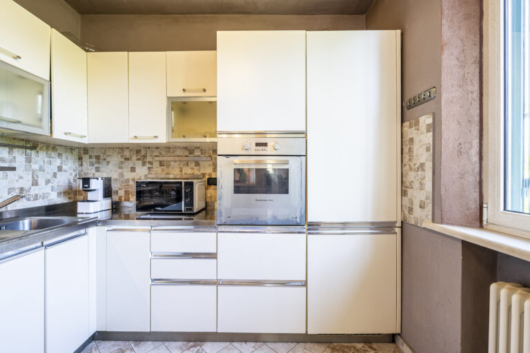 Cocina moderna con gabinetes blancos, horno empotrado, microondas, encimera de acero inoxidable y fondos de mosaico beige.